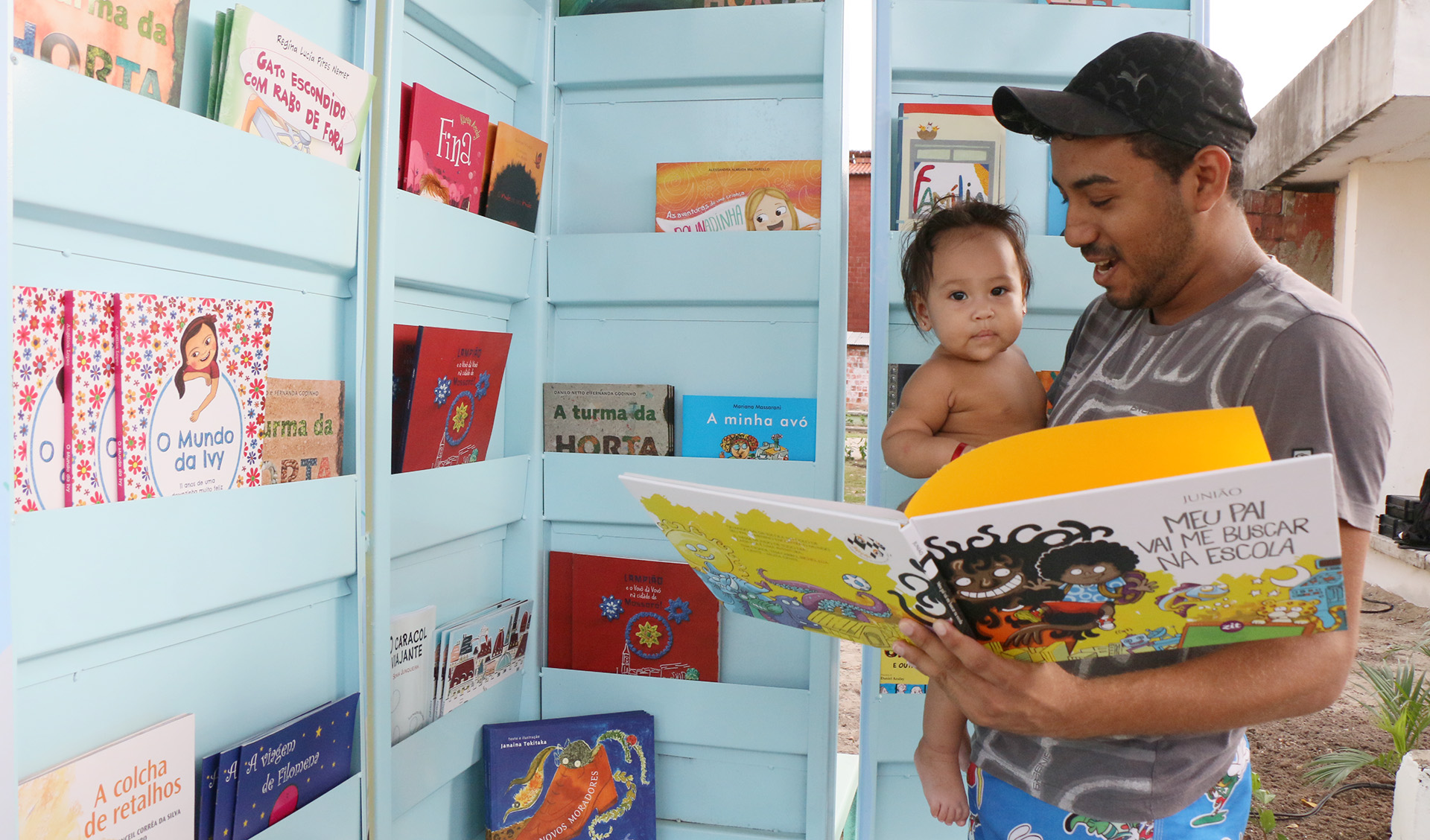 homem de boné segurando bebê no colo e livro infantil em frente a quiosque cheio de livros